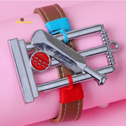 A bracelet with a cricket-themed charm is displayed against a pink background. The charm features a bat, ball, and stumps in a metallic finish, while the bracelet itself is brown leather with blue and red thread accents—an ideal rakhi for kids who love the game.