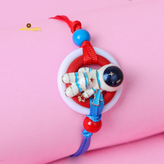 A colorful bracelet with a small astronaut charm lying on a pink surface, resembling a festive rakhi for kids. The bracelet features red and blue strings with round beads, and the astronaut is white with blue and red details, encircled by a red and white ring.