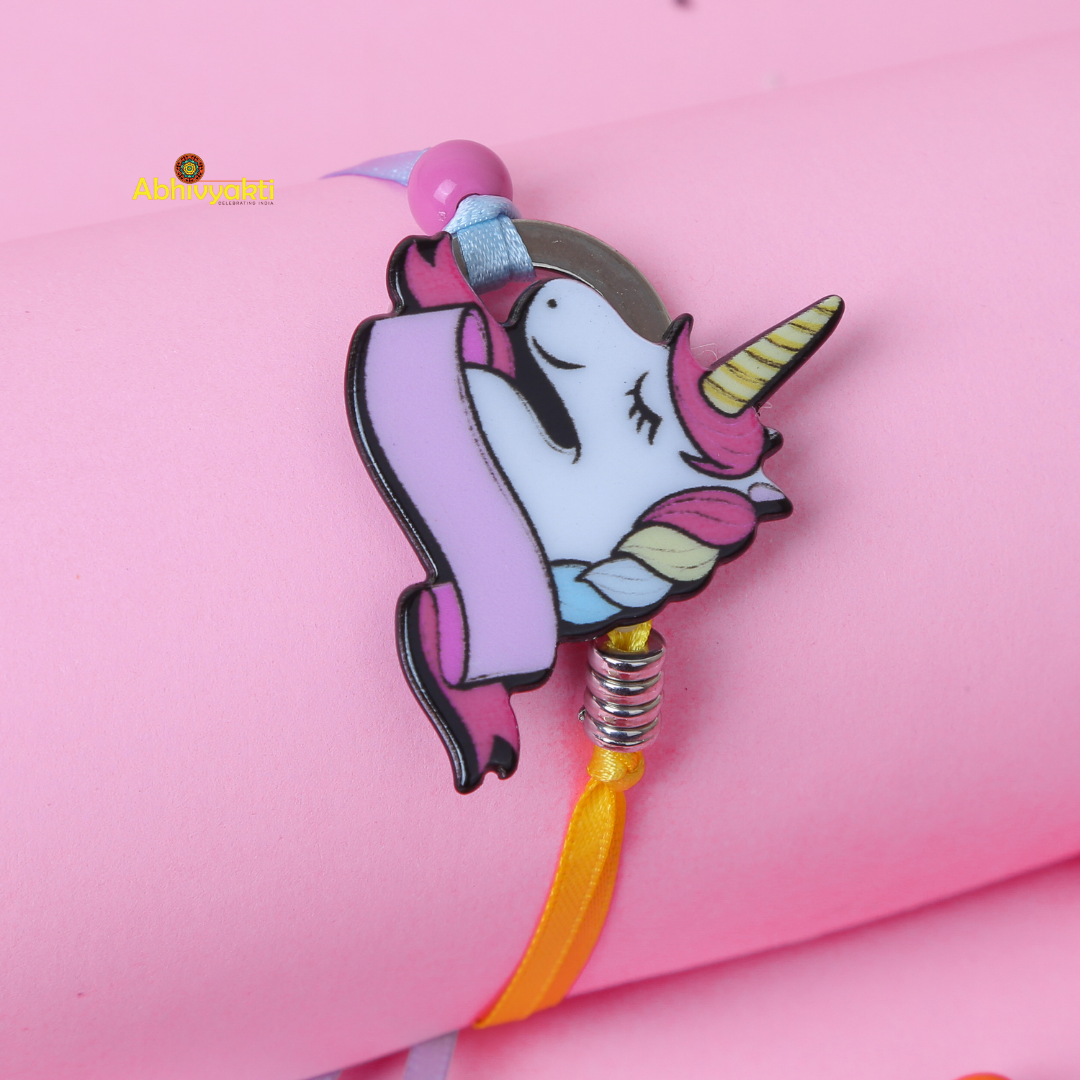A pink wristband with a cute unicorn charm featuring a pastel-colored mane and horn. The unicorn, smiling with closed eyes, is adorned with a decorative banner. Perfect as a rakhi for kids, the wristband also has a yellow ribbon and metallic spring detail against its pink background.