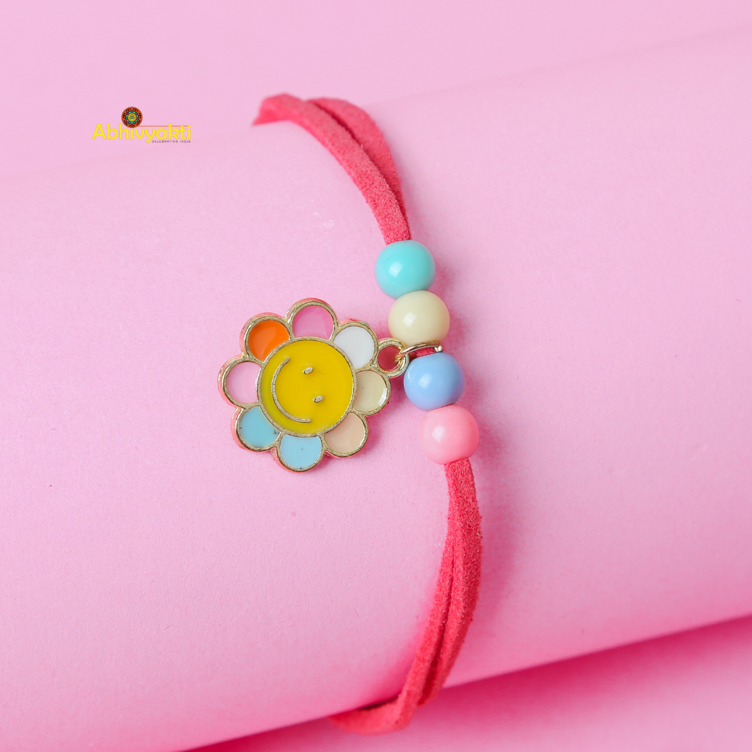 A pink bracelet with a smiling flower charm and colorful beads, perfect as a rakhi for kids, rests on a pink cylindrical object. The flower charm has petals in various colors, including yellow, orange, and blue, and the beads are in pastel shades of blue, yellow, and pink.