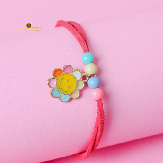 A pink bracelet with a smiling flower charm and colorful beads, perfect as a rakhi for kids, rests on a pink cylindrical object. The flower charm has petals in various colors, including yellow, orange, and blue, and the beads are in pastel shades of blue, yellow, and pink.