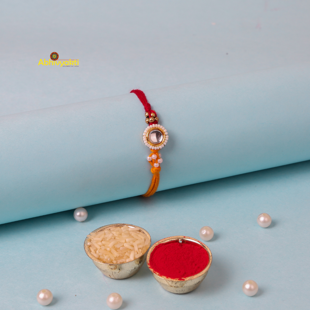 A close-up of a decorative kundan rakhi with stone and beads, featuring a central round gem surrounded by small white beads. It's tied on a red and orange thread with additional small beads.