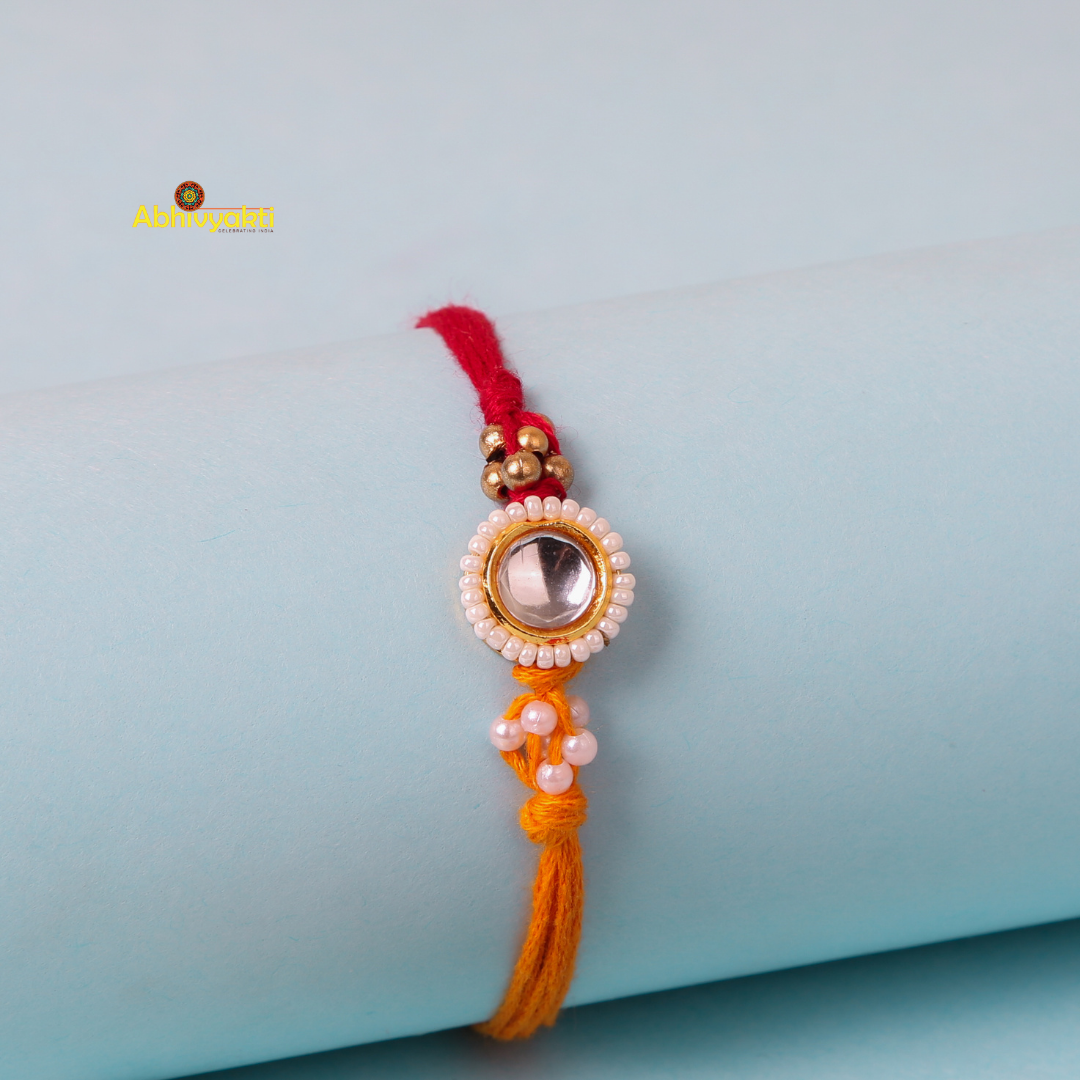 A close-up of a decorative kundan rakhi with stone and beads, featuring a central round gem surrounded by small white beads. It's tied on a red and orange thread with additional small beads. The rakhi is placed on a light blue cylindrical surface in the background.