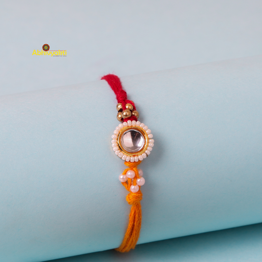 A close-up of a decorative kundan rakhi with stone and beads, featuring a central round gem surrounded by small white beads. It's tied on a red and orange thread with additional small beads. The rakhi is placed on a light blue cylindrical surface in the background.