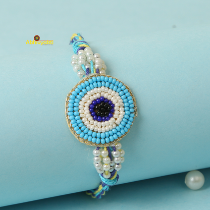 A close-up of a beaded bracelet featuring an eye motif made from concentric circles of beads in Sky blue, light blue, black, and white hues. The evil eye rakhi is displayed on a light blue cylindrical surface with a blue string band.