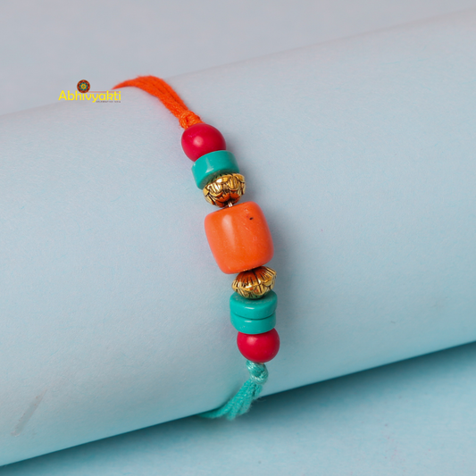 A colorful bracelet with orange and turquoise beads, along with gold spacers, is displayed on a light blue cylindrical object. The rakhi with beads features an orange thread on the left end and a turquoise thread on the right end.