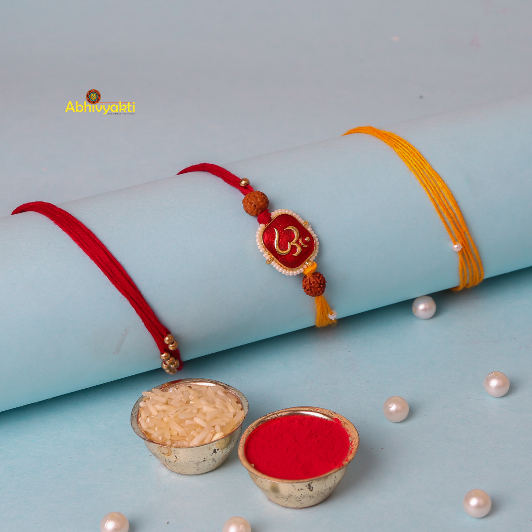 A rolled-up light blue sheet with two Rakhi bands, one red with a decorative 'Om' symbol and another yellow, featuring an Om Rakhi with long thread and rudraksha beads. Small bowls containing rice and red powder (kumkum) are in the foreground, with scattered pearls around them.