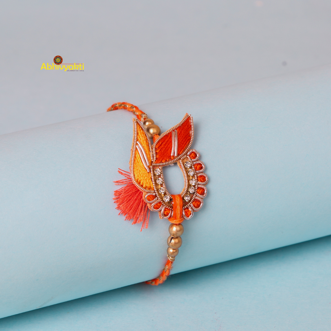 An artistic, orange and gold zari rakhi with beads and stone gracefully rests on a light blue cylindrical surface. The intricate design features leaf-like motifs, bead accents, and a central circular section adorned with small diamonds. A tassel detail adds a touch of elegance.