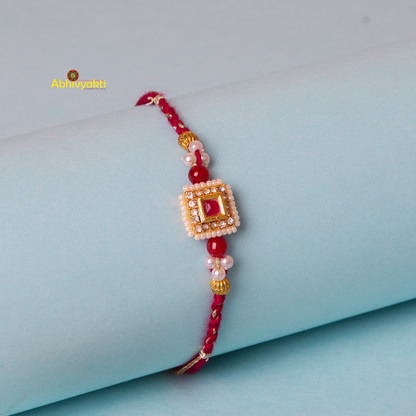A traditional stone rakhi with a red thread, adorned with a gold square centerpiece featuring small white beads and a red jewel in the center. The rakhi is wrapped around a light blue cylindrical object.