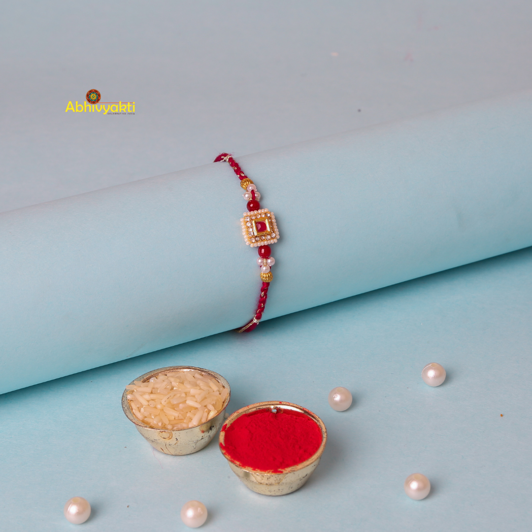 A traditional stone rakhi with a red thread, adorned with a gold square centerpiece featuring small white beads and a red jewel in the center