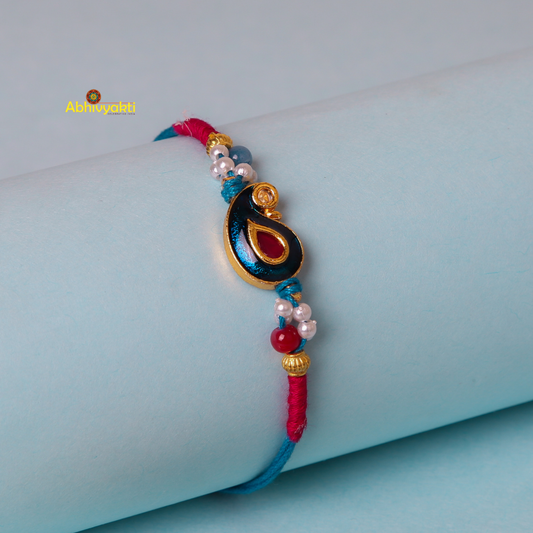 A decorative bracelet with red and blue threads wrapped around a cylindrical light blue object. The centerpiece is a gold emblem featuring a blue teardrop shape with a red inset, reminiscent of the intricate designs found in kundan paisley rakhi, surrounded by small beads in various colors.