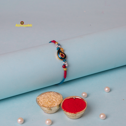 A decorative bracelet with red and blue threads wrapped around a cylindrical light blue object. The centerpiece is a gold emblem featuring a blue teardrop shape with a red inset, reminiscent of the intricate designs found in kundan paisley rakhi, surrounded by small beads in various colors.