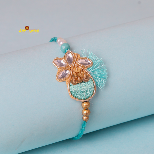 A delicate turquoise and gold stone rakhi with stone embellishments rests on a light blue cylindrical surface. The rakhi features intricate beadwork, golden floral designs, and mirror accents with tiny turquoise beads enhancing its elegance and a central golden bead cluster.