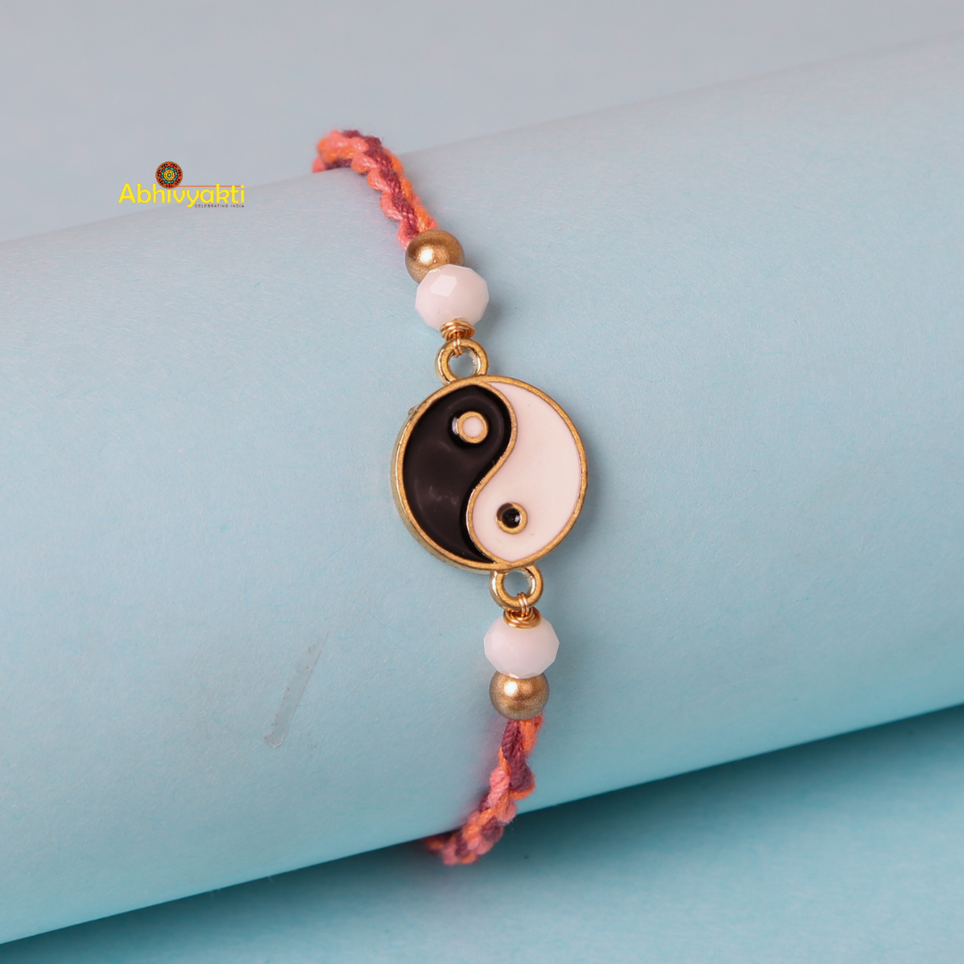 A bracelet with a Yin-Yang charm rests on a light blue cylindrical surface. The charm, circular with a gold rim, displays the classic black and white yin-yang design. It is attached to a braided multi-colored string with white and gold beads on either side, resembling an elegant enamel gold plated rakhi with stone and beads.