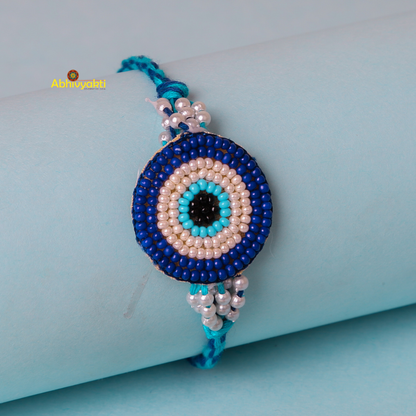 A close-up of a beaded bracelet featuring an eye motif made from concentric circles of beads in Dark blue, light blue, black, and white hues. The evil eye rakhi is displayed on a light blue cylindrical surface with a blue string band.