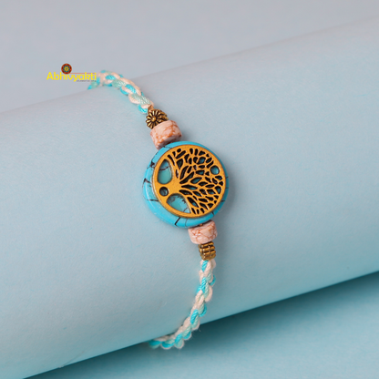 A braided bracelet with blue and white threads, featuring a circular charm with an intricate tree of life design in gold. The charm is flanked by two smaller pink beads and metallic accents, set against a pastel blue background—perfect as a gold plated rakhi for special occasions.