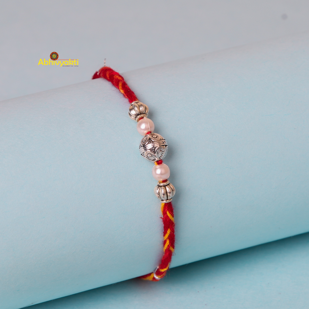 A close-up image of a rakhi with silver beads, braided with yellow and red thread and adorned with pink beads. The bracelet is wrapped around a light blue cylindrical object, displayed against a light blue background.