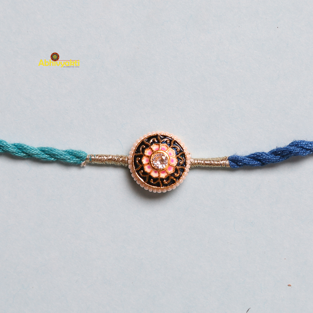 A close-up of a blue and teal braided rakhi with beads and stone, featuring a circular, gold-toned centerpiece. The centerpiece boasts an intricate floral design with a pink gemstone in the center, set against a light background.