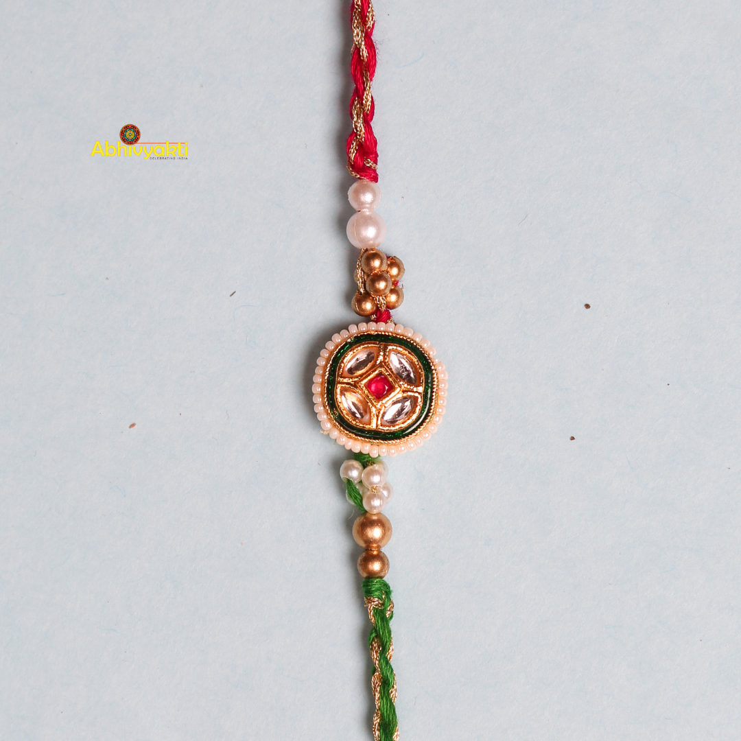 A traditional Rakhi featuring a central round design with red and green accents, surrounded by small pearls, intricate golden embellishments, and stone beads. The thread is braided in red and green, symbolizing the festival of Raksha Bandhan.