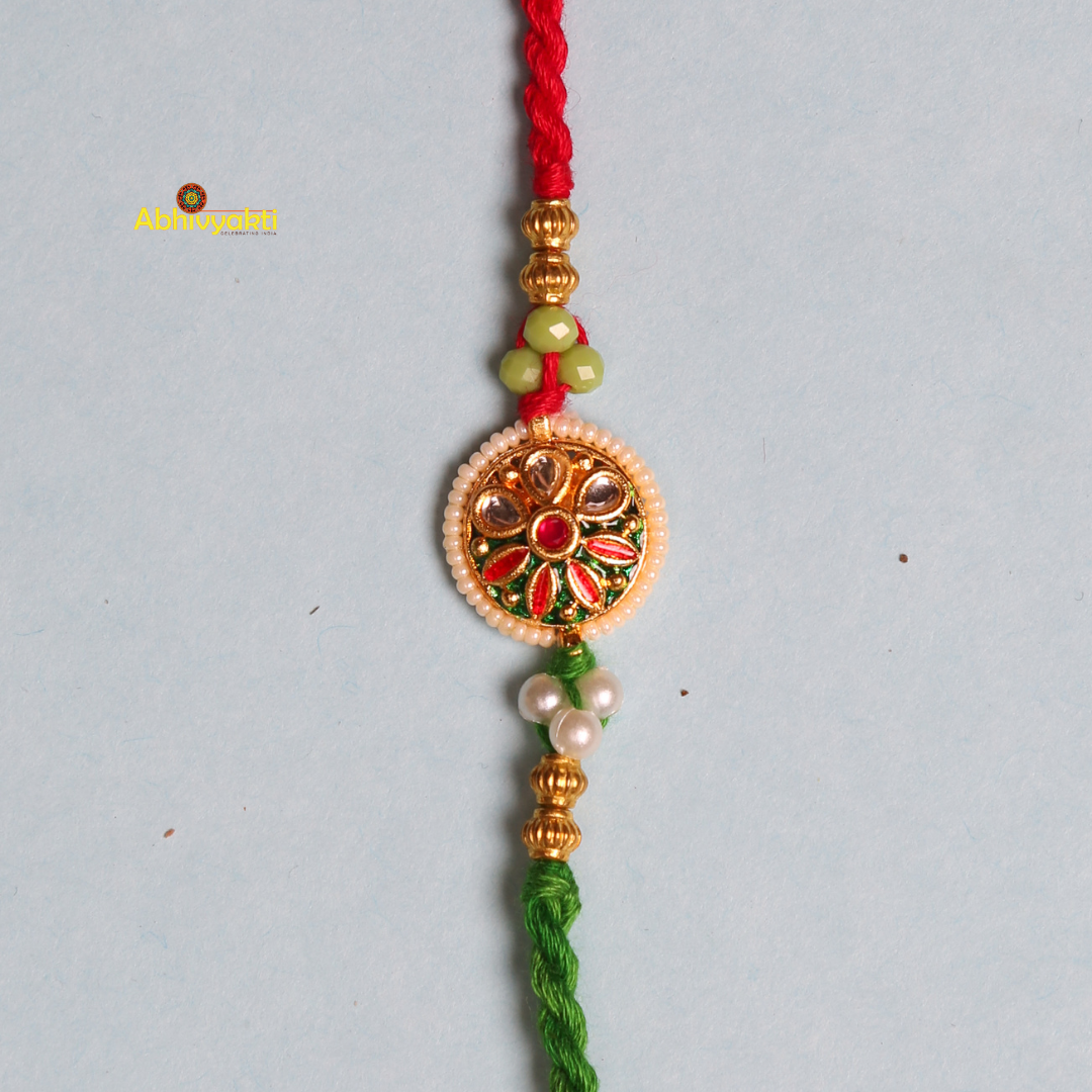 An ornate premium rakhi with a braided red and green thread and golden beads. The center pendant features a colorful, intricate multicolor floral kundan design with green, red, and pearl-like details against a light blue background.