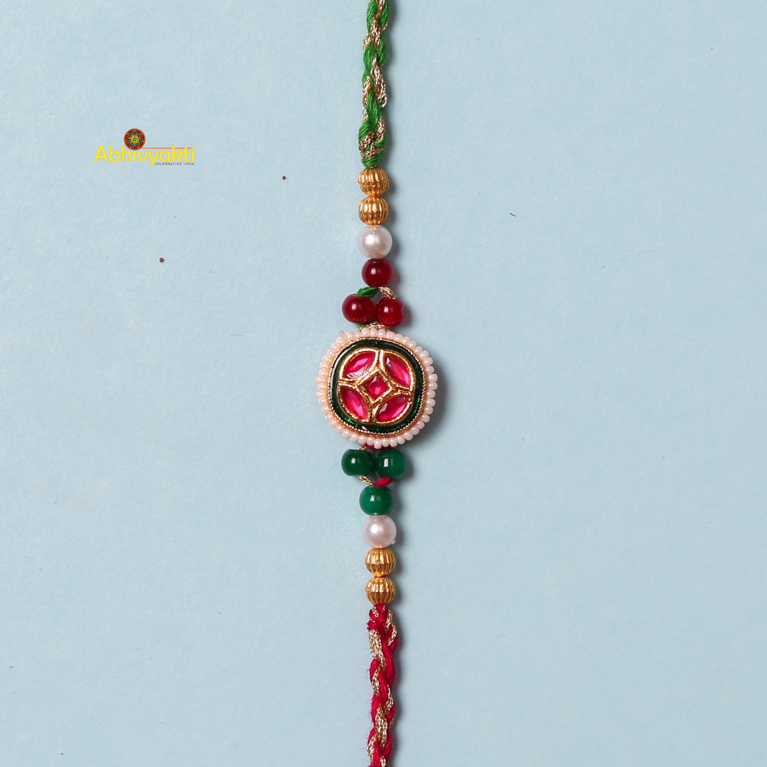 A beaded red and gold-plated rakhi against a light blue background. The rakhi features a central circular emblem with red and green intricate designs, flanked by beads of various colors including white, green, and gold, with a braided red and green thread.