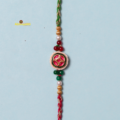 A beaded red and gold-plated rakhi against a light blue background. The rakhi features a central circular emblem with red and green intricate designs, flanked by beads of various colors including white, green, and gold, with a braided red and green thread.