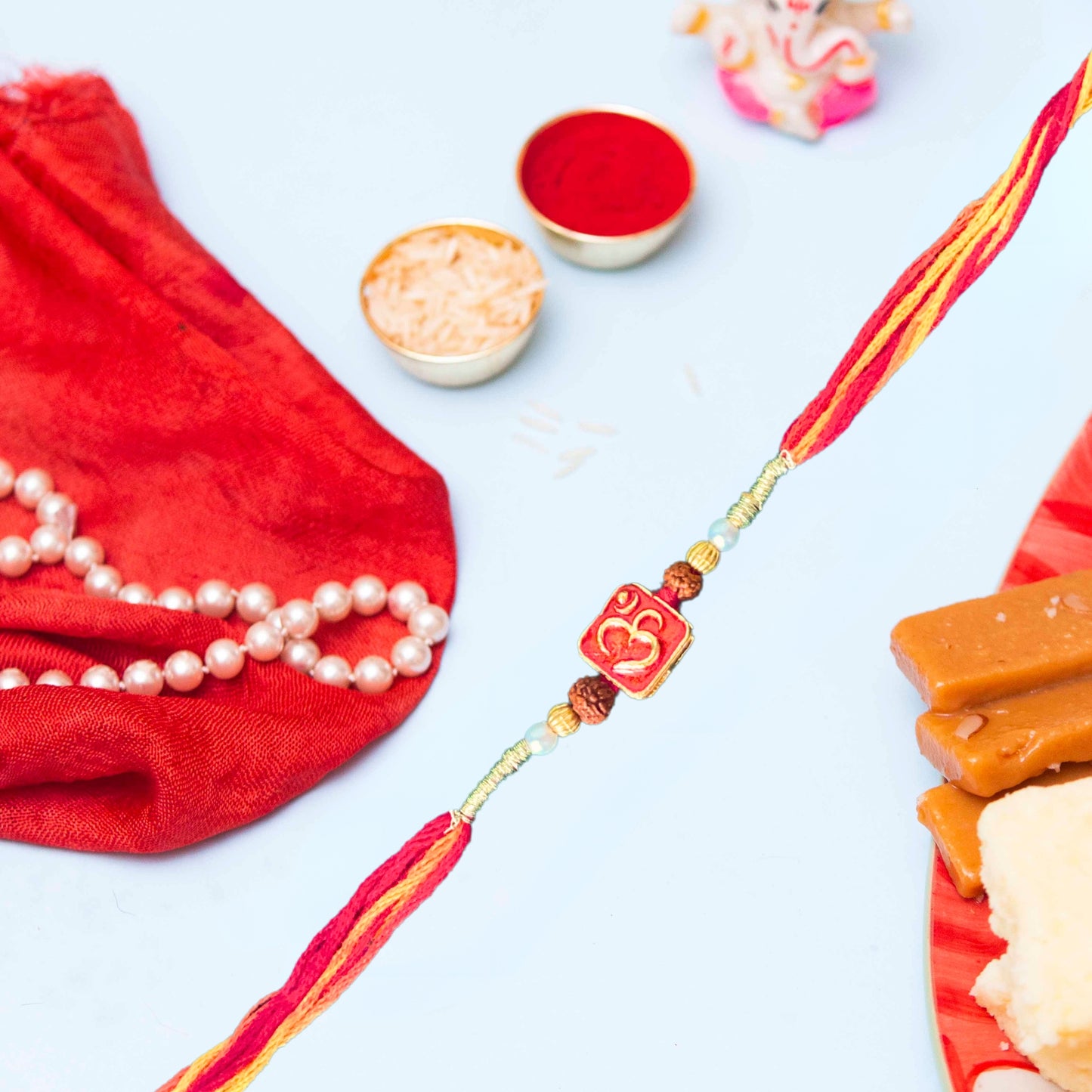 om rakhi with beads, showcasing a red and yellow thread band adorned with golden and rudraksha beads, features a red square centerpiece with a gold "Om" symbol, displayed against a pastel blue background