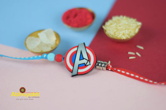A kids Rakhi with the Avengers logo and colorful beads, next to a bowl of rice.
