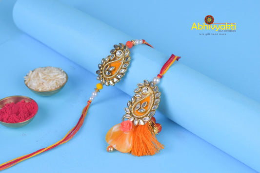Unique design orange rakhi and paisley lumba for bhaiya bhabhi with gold thread, tassels, stone, and beads.