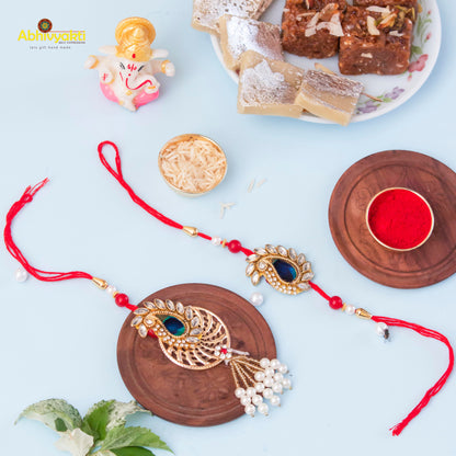 Pair of rakhi in gold and red thread, embellished with stones & beads, next to a plate of sweets.