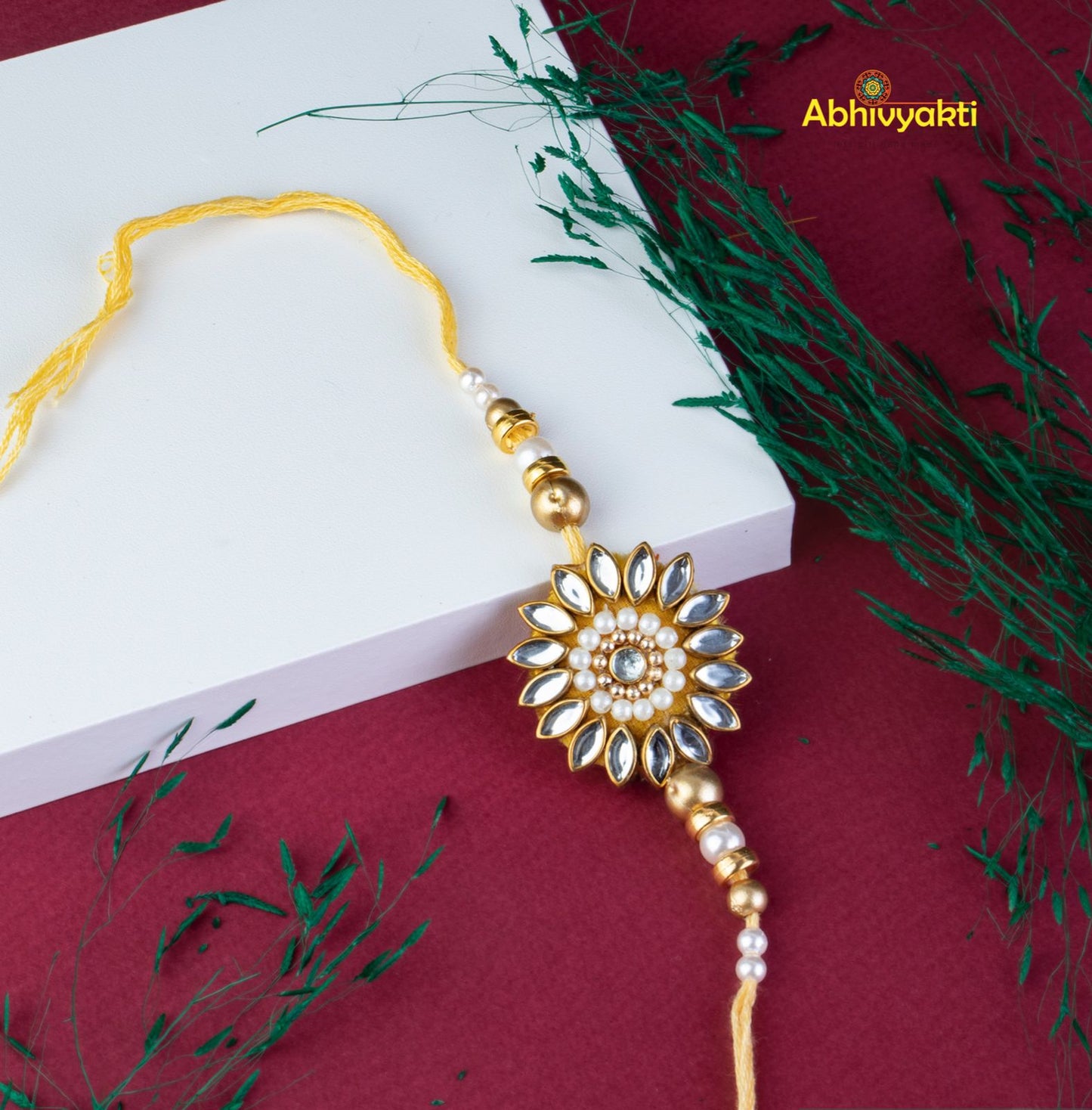 A gold and white rakhi with a flower, adorned with kundan stones and beautiful beads.